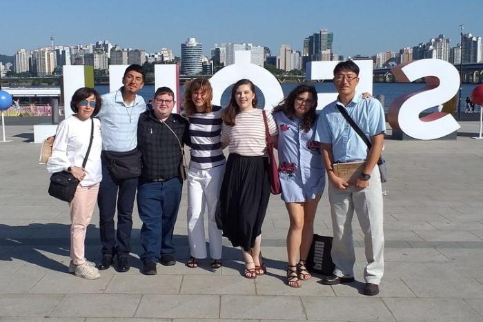 A group of seven people stands together, smiling towards the camera. Behind them, large white letters spell out "SEOUL". The backdrop features a river and a modern city skyline with numerous high-rise buildings under a clear blue sky.