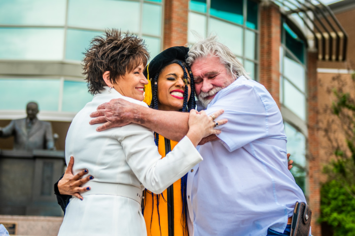 Three people are celebrating outdoors. A graduate in a yellow gown and black cap is embraced by an older man with white hair and a woman with short hair, both smiling. They stand in front of a modern building with large windows.