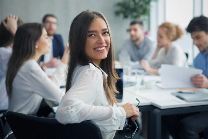 A woman with long brown hair and a white blouse is smiling at the camera in a modern office meeting room with several other people behind her.